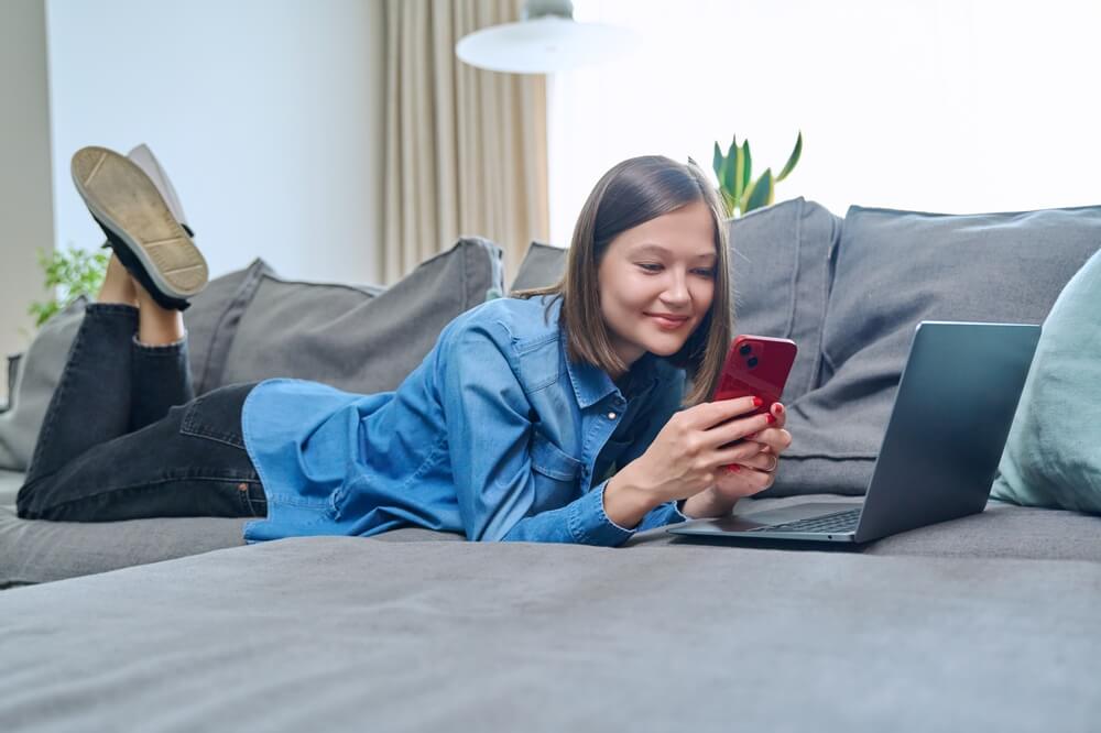 jente smiler og ligger på sofaen hjemme med mobilen i hånden.