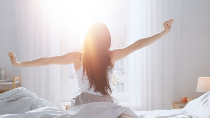 Kvinne sitter i seng og strekker armene mot morgenlyset som glipper inn mellom gardinene på soverommet.