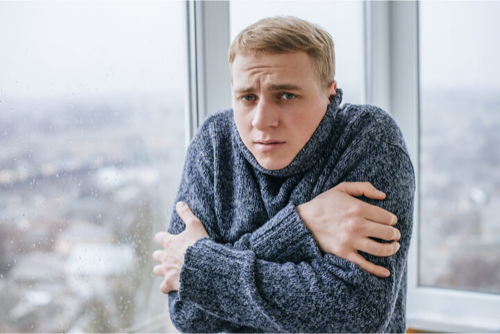 Kaldras oppleves som et kaldt trekk langs gulvet når temperaturen ute er kjølig. Her kan du finne ut hvorfor den oppstår og hva du kan gjøre for å bli kvitt det. Mann ved et stort, kaldt vindu hutrer og hakker tenner